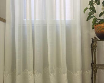 Może przedstawiać: Sięgająca podłogi, półprzezroczysta zasłona w kolorze złamanej bieli z pełnym pasem materiału u dołu. Dolna krawędź ozdobiona jest powtarzającym się haftowanym wzorem w kształcie zawijasów. Zasłona układa się w pionowe fałdy, przepuszczając światło.
