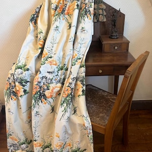 Op de afbeelding: Een wit stoffen gordijn met een bloemenprint met oranje, groene en zwarte bloemen. Het gordijn is gedrapeerd over een houten stoel en bureau.