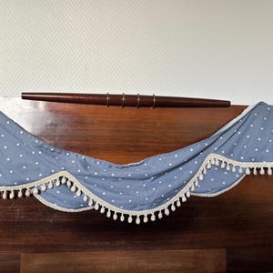 Op de afbeelding: Een blauw-witte polka dot valance met een crèmekleurige rand en kwastjes. De valance is gedrapeerd over een donker houten hoofdbord, met een donkere houten stang erboven.