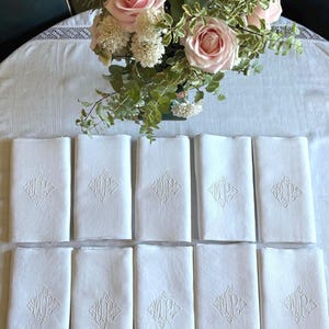 May include: A collection of ten white linen napkins, each embroidered with a monogram within a diamond shape. A bouquet of pink roses and greenery sits in the background on a white tablecloth. The napkins are neatly arranged.