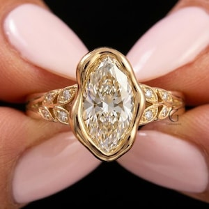 May include: A gold ring featuring a central marquise-cut gemstone surrounded by a smooth, organic-shaped gold bezel. The band displays a delicate leaf-like pattern on both sides, accented with small, round, clear inset stones along the metal shank.