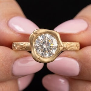 May include: A gold ring featuring a round, brilliant-cut clear gemstone set within an organic, wavy-edged bezel. The band has a matte finish and is engraved with small star and crescent moon motifs radiating from the central setting.