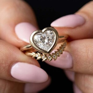 May include: A gold ring set featuring a heart-shaped center stone in a bezel setting paired with a curved band adorned with small, leaf-shaped accents and embedded stones. The two rings are stacked together to create a cohesive jewelry piece.