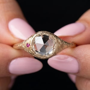 May include: A gold ring featuring a large, clear, faceted gemstone. A small, red gemstone is set to the side. The ring has an ornate, engraved design around the main stone and on the band.