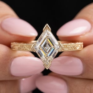 May include: A gold ring featuring a kite-shaped center stone held in a bezel setting. The band is crafted from yellow gold and displays an intricate, engraved scrollwork pattern along the sides, providing a detailed texture to the metal surface.