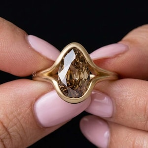 May include: A gold ring featuring a pear-shaped brown gemstone set in a smooth bezel setting. The band has a split design that curves around the central stone, creating a continuous frame. The metal has a matte finish and a warm yellow tone.