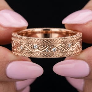May include: A rose gold band ring featuring an intricate engraved scrollwork pattern across the centre, bordered by two rows of milgrain detailing. Several small, round, clear white diamonds are set within the swirling engraved design.