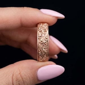 May include: A rose gold band ring featuring an intricate, raised floral and leaf scrollwork pattern engraved across the entire outer surface. The metal has a warm, metallic pink hue with polished highlights that accentuate the detailed carvings.