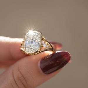 Op de afbeelding: Een gouden ring met een grote, rechthoekig geslepen diamant als middelpunt. De ring heeft een gedetailleerde zetting met kleinere accentdiamanten en een vintage-geïnspireerd ontwerp. De ring wordt tegen een vage achtergrond gehouden.