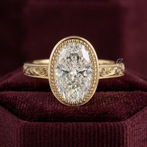 May include: A gold ring featuring a large oval-cut center stone surrounded by a beaded bezel setting. The band is crafted from yellow gold and displays intricate, engraved scrollwork patterns along the sides, extending from the central setting.