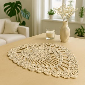 May include: A beige, fan-shaped, crocheted doily sits on a light-coloured wooden table. The doily has an intricate openwork design. A small candle in a glass holder and a cream-coloured vase with dried flowers are also on the table. A white sofa and plants are in the background.