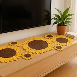 Puede incluir: Un conjunto de decoración de mesa de girasoles hechos a mano con ganchillo. El conjunto incluye un camino de mesa y varios posavasos a juego. Los girasoles tienen pétalos amarillos y centros marrones.