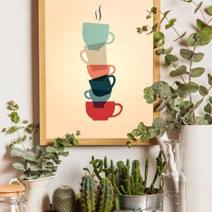 Op de afbeelding: Een ingelijst afdruk van een stapel kleurrijke koffiekopjes met stoom die uit het bovenste kopje opstijgt. De afdruk hangt aan een witte muur met planten en vetplanten in potten op de voorgrond.
