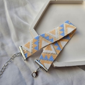 May include: A beaded bracelet with a geometric pattern in white, light blue, and tan. The bracelet has silver-colored metal clasps and a chain for adjustable sizing. The bracelet is on a white surface.
