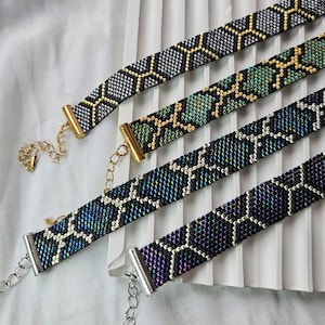 May include: Four beaded choker necklaces with geometric patterns. The chokers have gold, silver, and black hardware. The beadwork includes black, silver, green, and iridescent beads. The necklaces are displayed on a white surface.