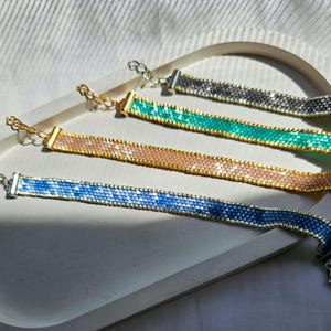 Peut inclure: Quatre bracelets plats en perles avec un motif tissé en grille dans les tons bleu, pêche, sarcelle et gris. Chaque bracelet est doté d'une finition en métal doré, incluant un fermoir mousqueton et une chaînette d'extension réglable.