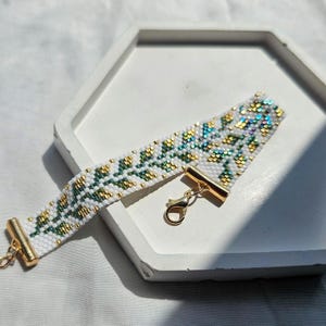 May include: A woven bead bracelet featuring a white background with a repeating leaf pattern in shades of green, gold, and iridescent blue. The bracelet is finished with gold-toned metal end caps and a matching lobster claw clasp closure.