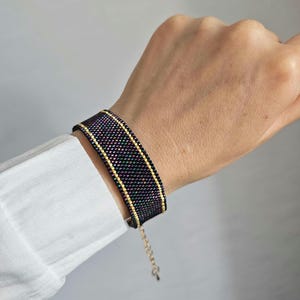 May include: A wide woven bead bracelet featuring a dark, iridescent purple and green central panel bordered by two thin rows of metallic gold beads. The bracelet includes a delicate gold-toned metal chain extension with a small circular clasp.
