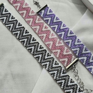May include: Four beaded bracelets in black, white, pink, and purple, with a zig-zag pattern. The bracelets have silver clasps. The bracelets are handmade. The measurements are in centimetres.