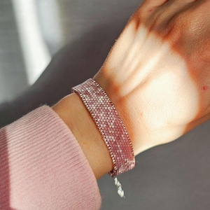 May include: A pink beaded bracelet with a delicate pattern is worn on a wrist. The bracelet is made of small, square beads in shades of pink and white, with a metallic border. The bracelet is approximately 0.75 inches wide.