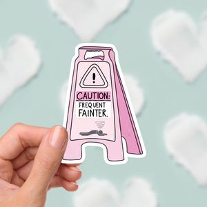 May include: A pink and white sticker in the shape of a caution sign. The sticker reads "CAUTION: FREQUENT FAINTER" with an image of a person lying down. The background is a light blue colour with white heart shapes.