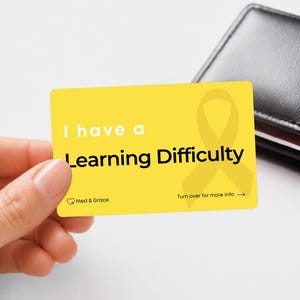 May include: A rectangular yellow card featuring the text I have a Learning Difficulty in black, a yellow awareness ribbon graphic, and the Med & Grace logo. The card includes the text Turn over for more info with an arrow pointing to the right.