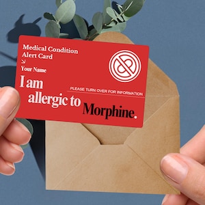May include: A red medical alert card with white text and a symbol, held in front of a brown envelope. The card reads "Medical Condition Alert Card" and "I am allergic to Morphine."