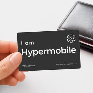 May include: A rectangular black medical alert card with white text that reads I am Hypermobile, featuring a white Star of Life symbol, a small heart icon with the text Med & Grace, and the instruction Turn over for more info with an arrow.