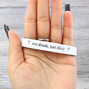 May include: A white bracelet with the text "not drunk, just dizzy" in black lettering. The bracelet also features a small medical symbol on each end. The bracelet is held in a hand.