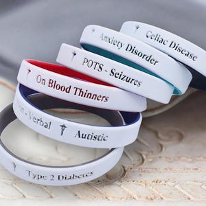May include: A stack of six white silicone wristbands with different medical alert messages, including "Celiac Disease", "Anxiety Disorder", "POTS - Seizures", "On Blood Thinners", "Autistic", and "Type 2 Diabetes".