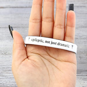 May include: White wristband with the text "epileptic, not just dramatic" in black. The wristband has a simple design with a small cross symbol on each end. The image shows the wristband on a hand.