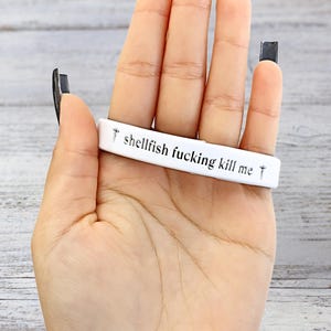 Peut inclure: Un bracelet blanc avec le texte "shellfish fucking kill me" en lettres noires. Le bracelet est tenu dans une main avec du vernis à ongles noir.