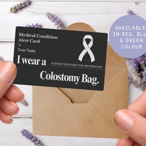 May include: A black medical alert card with white text and a white ribbon symbol. The card reads "I wear a Colostomy Bag." and "Medical Condition Alert Card." The card is held over a brown envelope. Available in red, blue, and green.