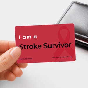 Peut inclure: Une carte d'alerte médicale rectangulaire rouge avec le texte blanc I am a et le texte noir Stroke Survivor. La carte présente un ruban de sensibilisation rouge, le logo Med & Grace et le texte Turn over for more info avec une flèche.