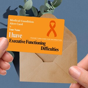 May include: An orange medical alert card with the text "Medical Condition Alert Card" and "I have Executive Functioning Difficulties." The card features a ribbon graphic and is held in front of a brown envelope.