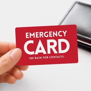 May include: A rectangular red emergency card with white text that reads EMERGENCY CARD and SEE BACK FOR CONTACTS. The card is held in a position that displays the front side clearly, with a black leather wallet visible in the background.