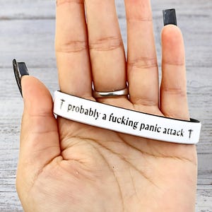 May include: White and black bracelet with the text "probably a fucking panic attack" in black. The bracelet is held in a hand with a silver ring and black nail polish.