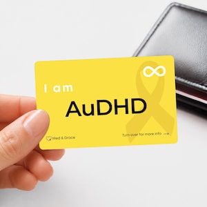 May include: A rectangular yellow card featuring the text I am AuDHD in bold black letters. The card includes a white infinity symbol, a yellow awareness ribbon graphic, and the text Med & Grace with a heart icon and Turn over for more info.