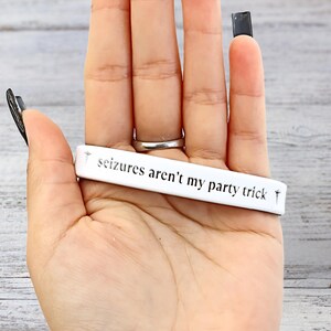 May include: A white bracelet with the text "seizures aren't my party trick" in black lettering. The bracelet is held in a hand with a silver ring and black nail polish.