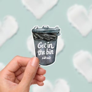 May include: A sticker depicting a grey watercolour illustration of a bin with the words "Get in the bin xoxo" in white. The sticker is held by a hand against a backdrop of white heart shapes on a light blue surface.
