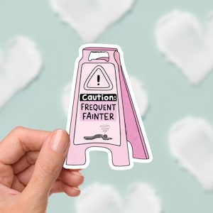 May include: A pink sticker in the shape of a hazard sign, with the text "Caution: Frequent Fainter." The sign features a graphic of a person lying down and a swirling symbol. The background is a light blue with white heart shapes.