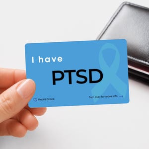 May include: A rectangular blue medical alert card featuring the white text I have PTSD, a large light blue awareness ribbon, and the small text Med & Grace with an arrow pointing right and the words Turn over for more info.