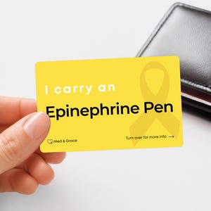 May include: A rectangular yellow medical alert card with rounded corners. The text reads I carry an Epinephrine Pen in white and black lettering. A yellow awareness ribbon graphic is on the right side. The Med & Grace logo and Turn over for more info text are visible.