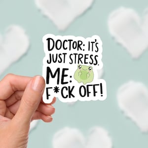 May include: A white sticker with black text that reads "DOCTOR: IT'S JUST STRESS. ME: F*CK OFF!" A small green frog illustration is in the center. The sticker is held by a hand against a light blue background with white heart shapes.