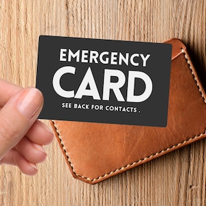 May include: A black emergency card with white text is held near a brown leather wallet. The card reads "EMERGENCY CARD" and "SEE BACK FOR CONTACTS." The wallet is rectangular with stitched edges, resting on a wooden surface.