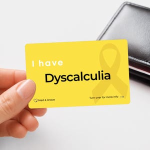 May include: A rectangular yellow card featuring the white text I have and the black text Dyscalculia. A yellow awareness ribbon is printed on the right side. The bottom left corner displays the text Med & Grace with a heart icon, and the bottom right corner says Turn over for more info with an arrow.