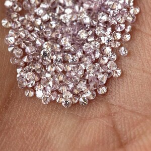 Puede incluir: Primer plano de una mano que sostiene una colección de pequeños diamantes rosa claro y brillantes. Los diamantes son de talla redonda y reflejan la luz, creando una exhibición brillante. Los diamantes están dispersos por la palma.