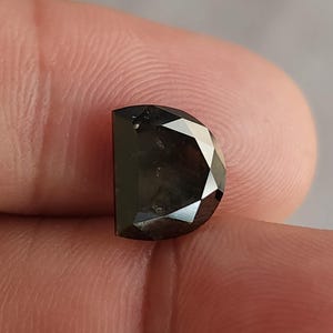 Könnte beinhalten: Ein facettierter, halbmondförmiger, schwarzer Diamant-Edelstein. Der Edelstein wird zwischen Fingern gehalten, wodurch seine polierten Oberflächen und dunkle Farbe zur Geltung kommen. Der Schliff des Diamanten erzeugt mehrere reflektierende Facetten.