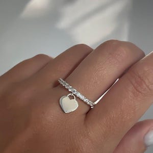 Puede incluir: Un anillo plateado con una banda de pequeñas piedras transparentes. Un pequeño dije de corazón pulido cuelga del anillo. El anillo está en un dedo.