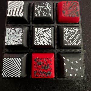 Könnte beinhalten: Ein Set aus neun quadratischen mechanischen Tastaturkappen, angeordnet in einem drei-mal-drei Raster. Die Tastenkappen haben schwarze oder rote Basen mit verschiedenen handgemalten abstrakten Mustern in Weiß oder Schwarz, darunter Linien.